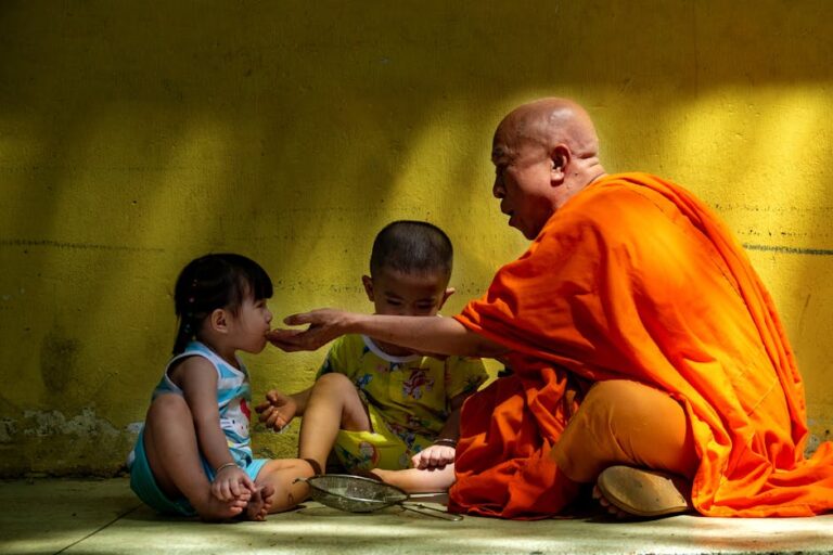 A Buddhist monk in orange robes feeds a young child against a yellow wall.