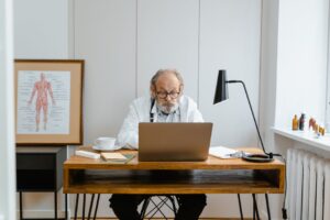 Elderly doctor in office using a laptop with medical books and anatomical chart.