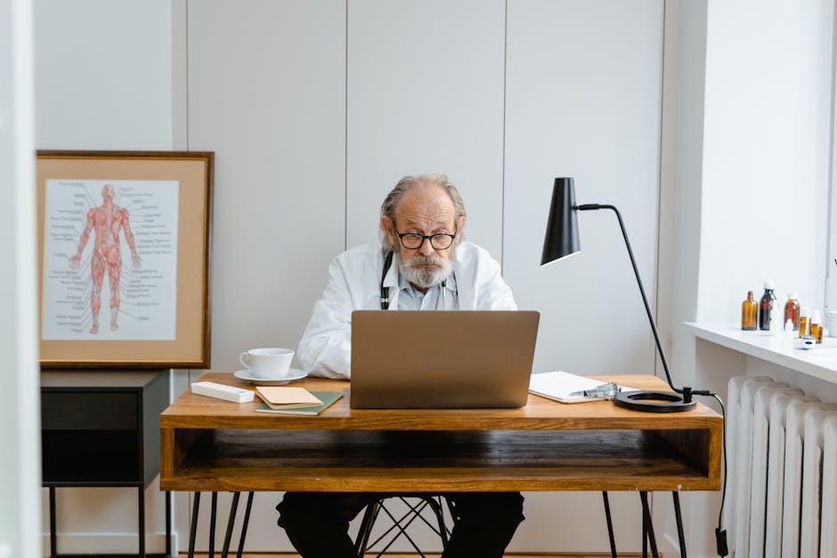 Elderly doctor in office using a laptop with medical books and anatomical chart.