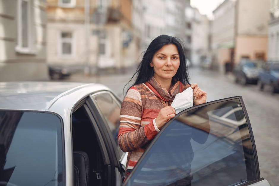 A woman stands beside an open car door in a city street, holding a protective face mask.