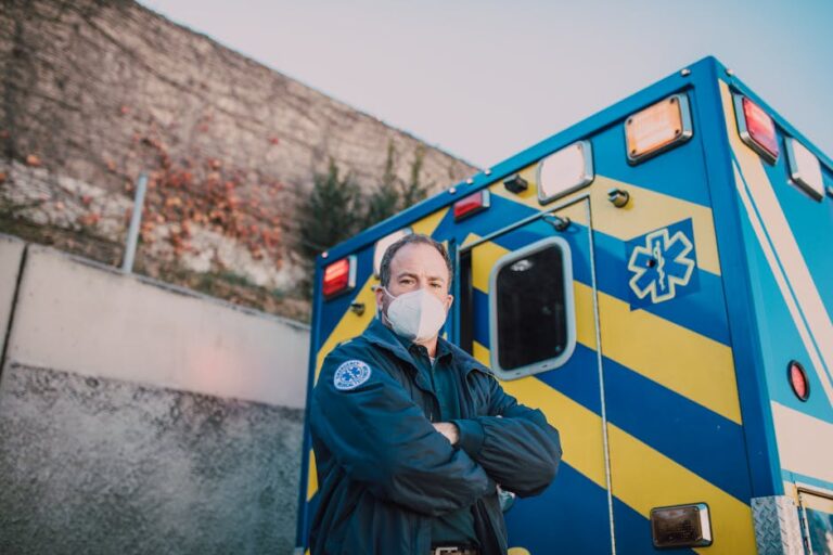 A confident paramedic wearing a mask stands arms crossed by an ambulance.