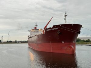 A large red cargo ship named Leonora Victory at a European dock with a wind turbine in the background.