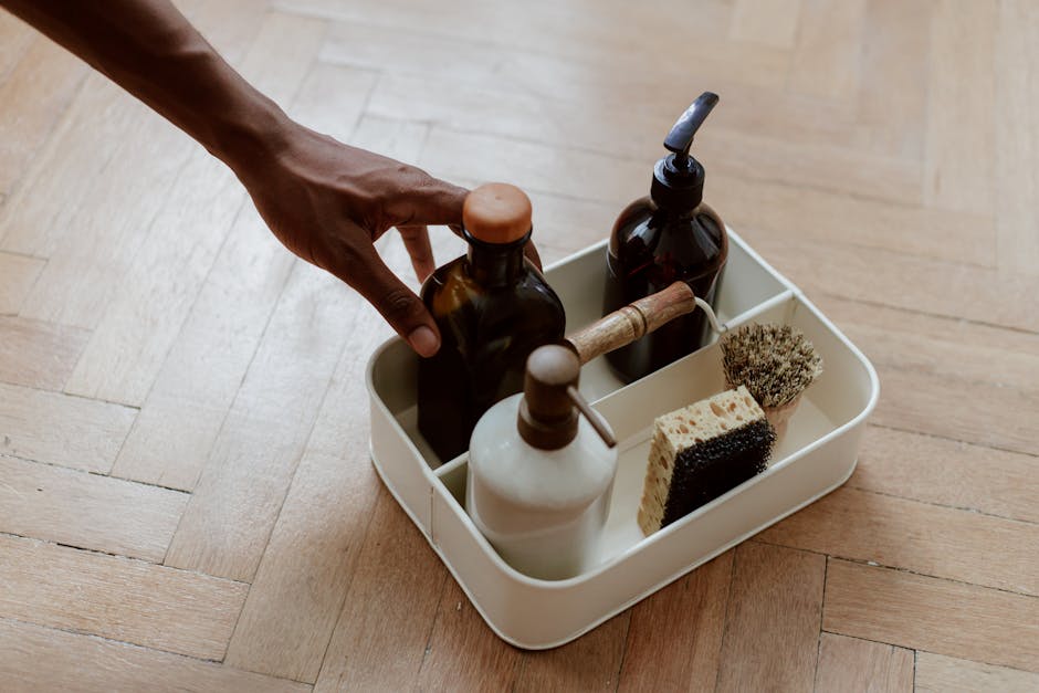A hand reaching for a cleaning set with bottles and brushes on a wooden floor.