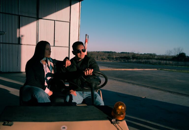 A couple sits in a small vehicle at an airport hangar during sunset, creating a serene scene.
